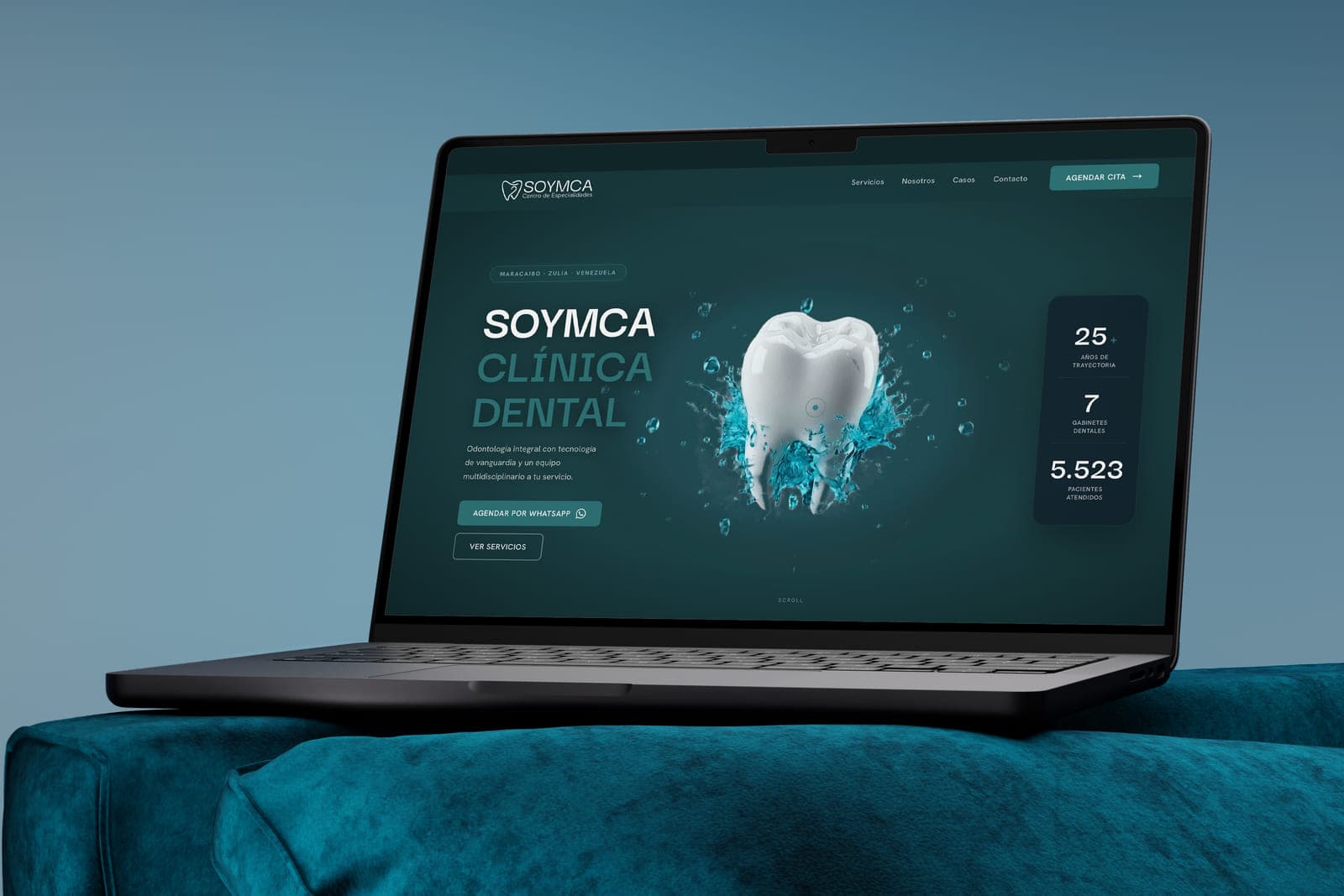 La home de Soymca en un portátil sobre un sofá azul petróleo: un diente en 3D rodeado de agua, el titular «Clínica Dental» y las cifras de la clínica al lado.
